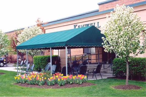 Outdoor seating area with a green canopy covering several black metal chairs and tables. The area is surrounded by blooming tulips and small flowering trees, with a building in the background.