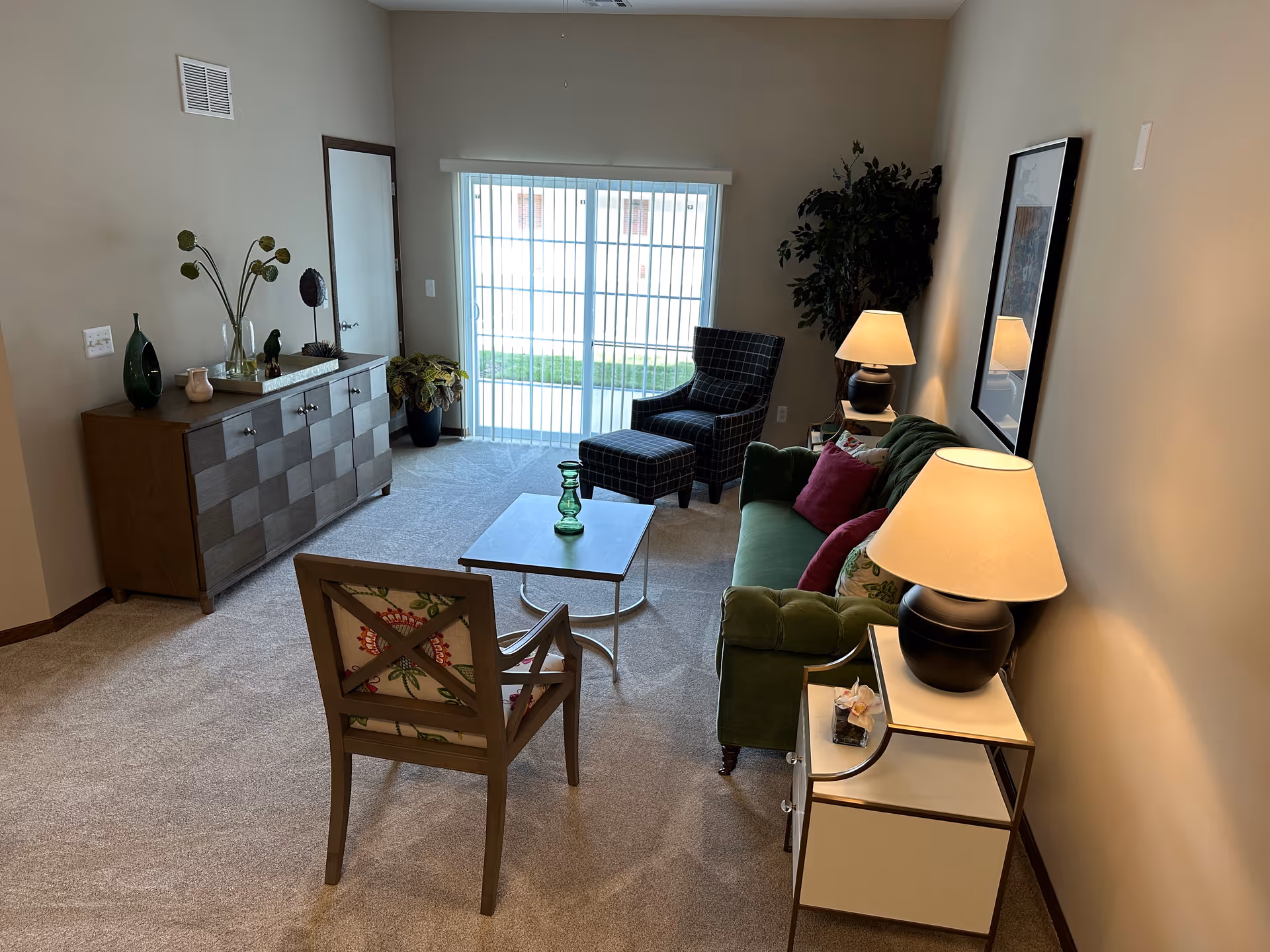 Cozy living room with a green sofa, armchair and ottoman, coffee table, sideboard and sliding glass door letting in natural light.