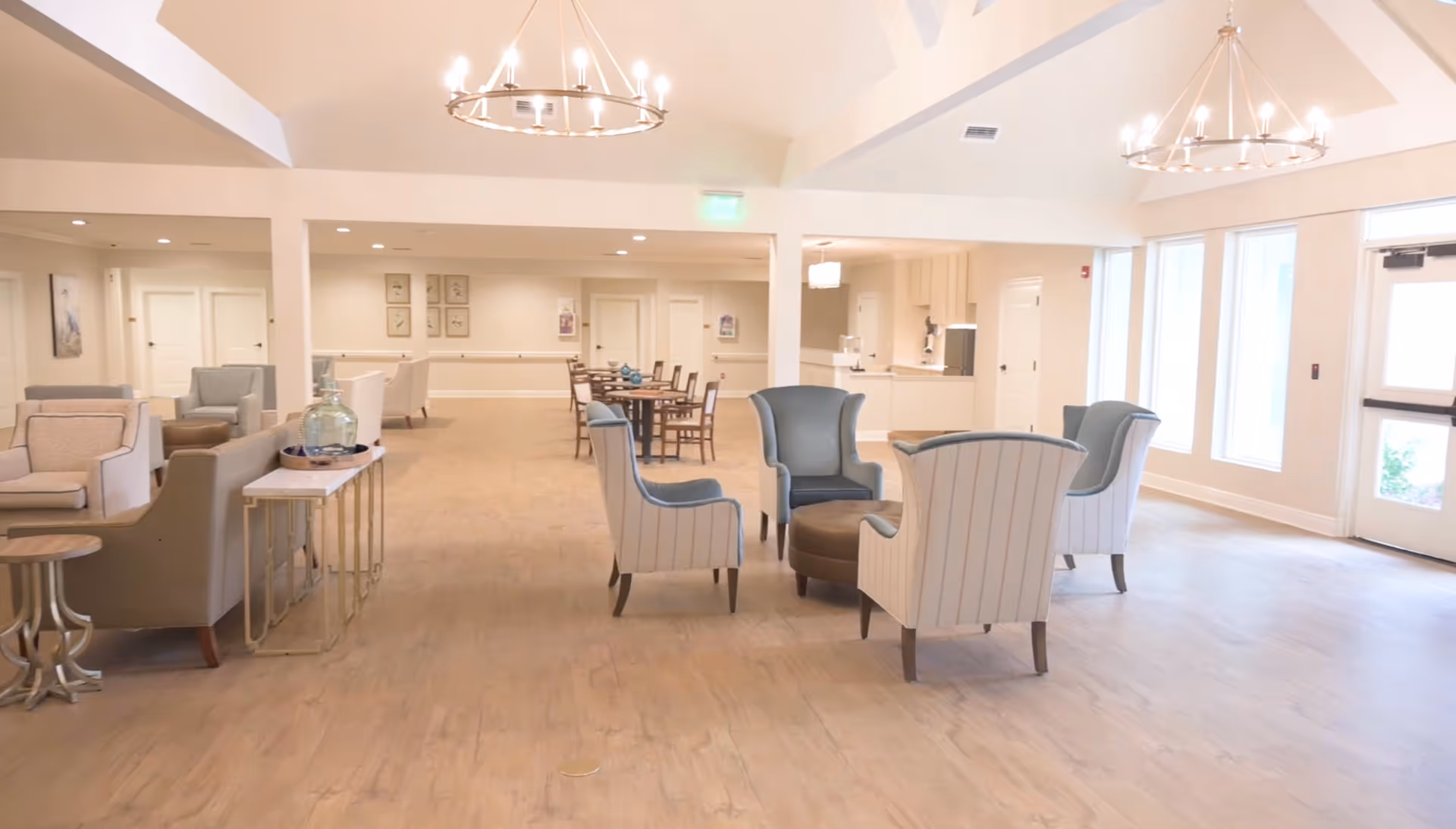 A spacious and well-lit common area in a senior living facility featuring several armchairs arranged around a round ottoman, a dining table with chairs in the background, light wood flooring, large windows letting in natural light, and modern chandeliers hanging from a high ceiling.