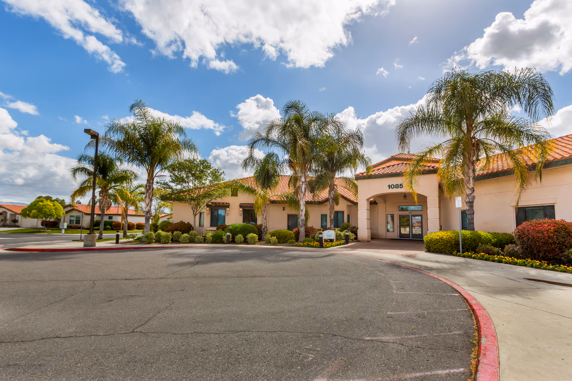 Exterior view of Brookdale Sunwest facility with a curved driveway, palm trees, and a beige building with a red-tiled roof under a partly cloudy sky.