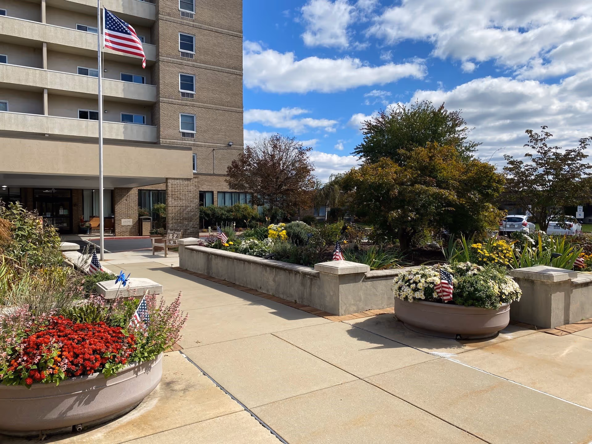 Outdoor view of Oak Hill Village showing the entrance area with a flagpole flying an American flag, large planters filled with colorful flowers and small American flags, a garden area with trees and shrubs, and part of the building with balconies and windows under a partly cloudy sky.