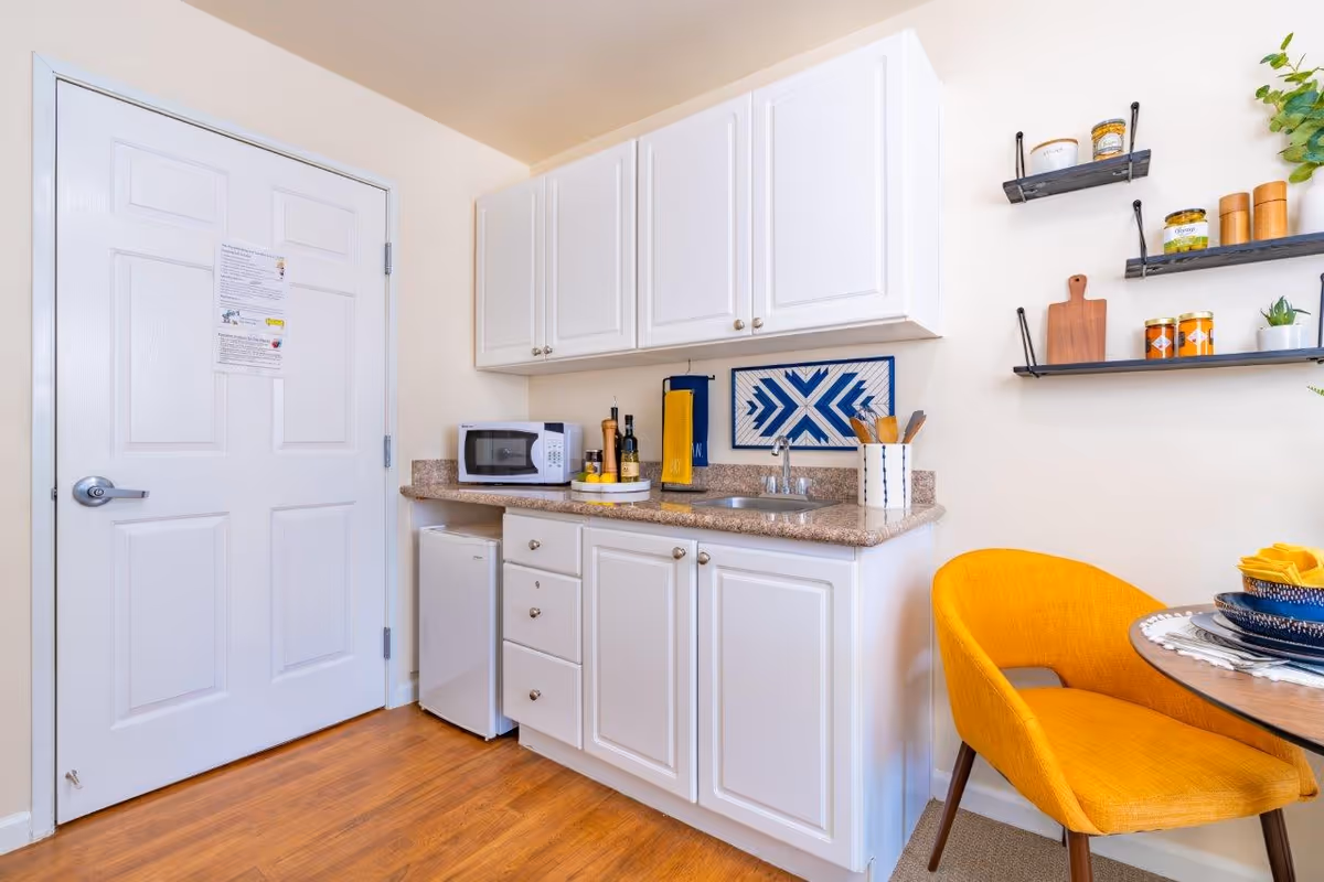 Small kitchenette with white cabinets, countertop sink, microwave and mini-fridge next to a yellow dining chair and table.