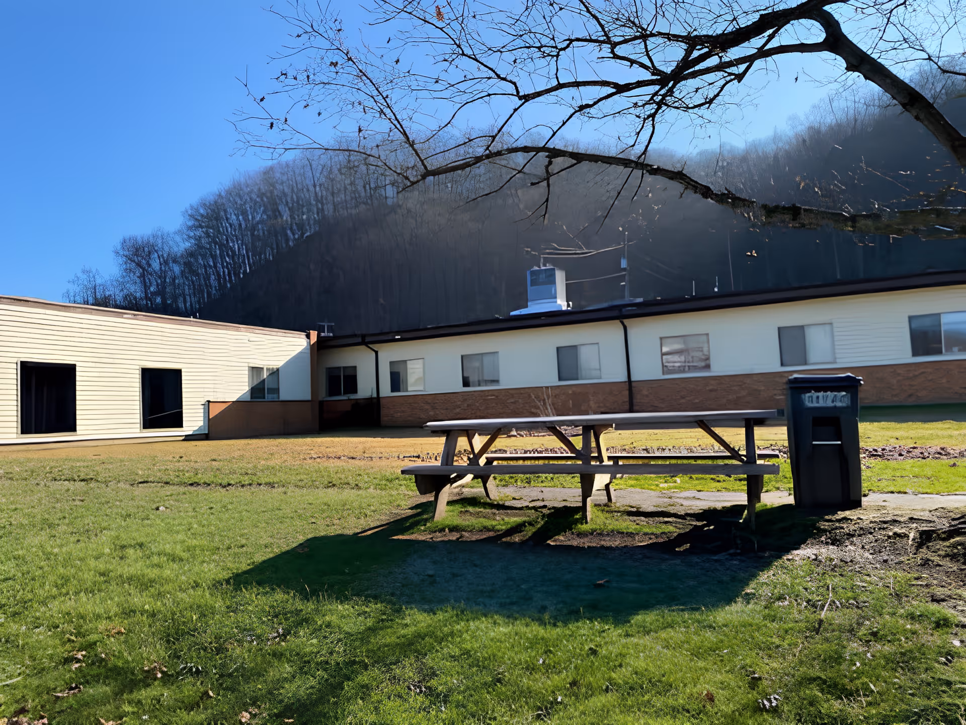 Picnic table and trash bin on a grassy courtyard in front of a single-story senior living building with wooded hills behind.