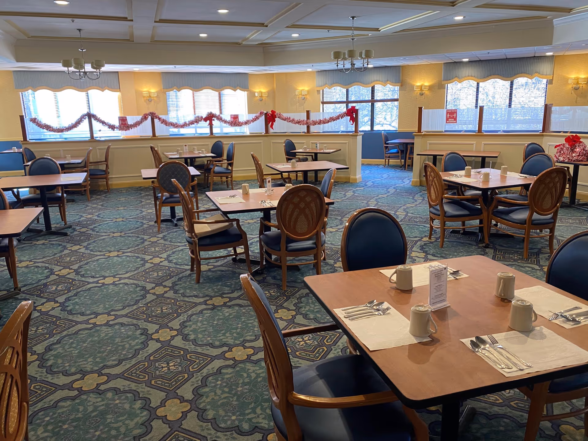 A dining room with multiple wooden tables and chairs arranged neatly. Each table is set with placemats, silverware, and white mugs. The room has large windows with blinds, decorated with red garlands and bows, allowing natural light to brighten the space. The carpet has a patterned design in shades of green and blue, and the walls are light-colored with sconces and chandeliers providing additional lighting.