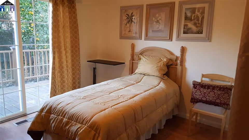 Small single bedroom with a twin bed, headboard, framed pictures, a sliding glass door to a deck, and a chair.