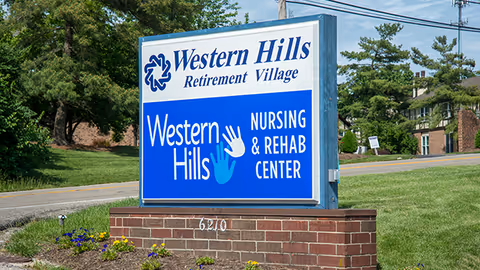 Outdoor sign for Western Hills Retirement Village and Western Hills Nursing & Rehab Center, situated on a brick base with grass and trees in the background along a roadside.