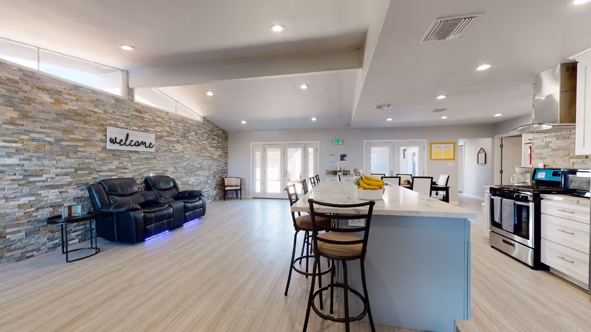 Open communal interior with a central marble island and bar stools, stainless kitchen appliances to the right, and a lounge area with black recliners against a stone accent wall displaying a 'welcome' sign.