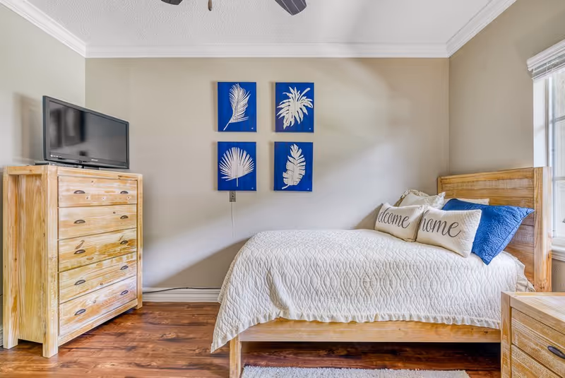 A cozy bedroom with a wooden bed frame and a white quilted bedspread. There are two decorative pillows on the bed, one with the word 'welcome' and the other with the word 'home'. On the wall above the bed are four blue square art pieces featuring white leaf designs. To the left of the bed is a wooden dresser with a flat-screen TV on top. The room has wooden flooring and a window on the right side letting in natural light.