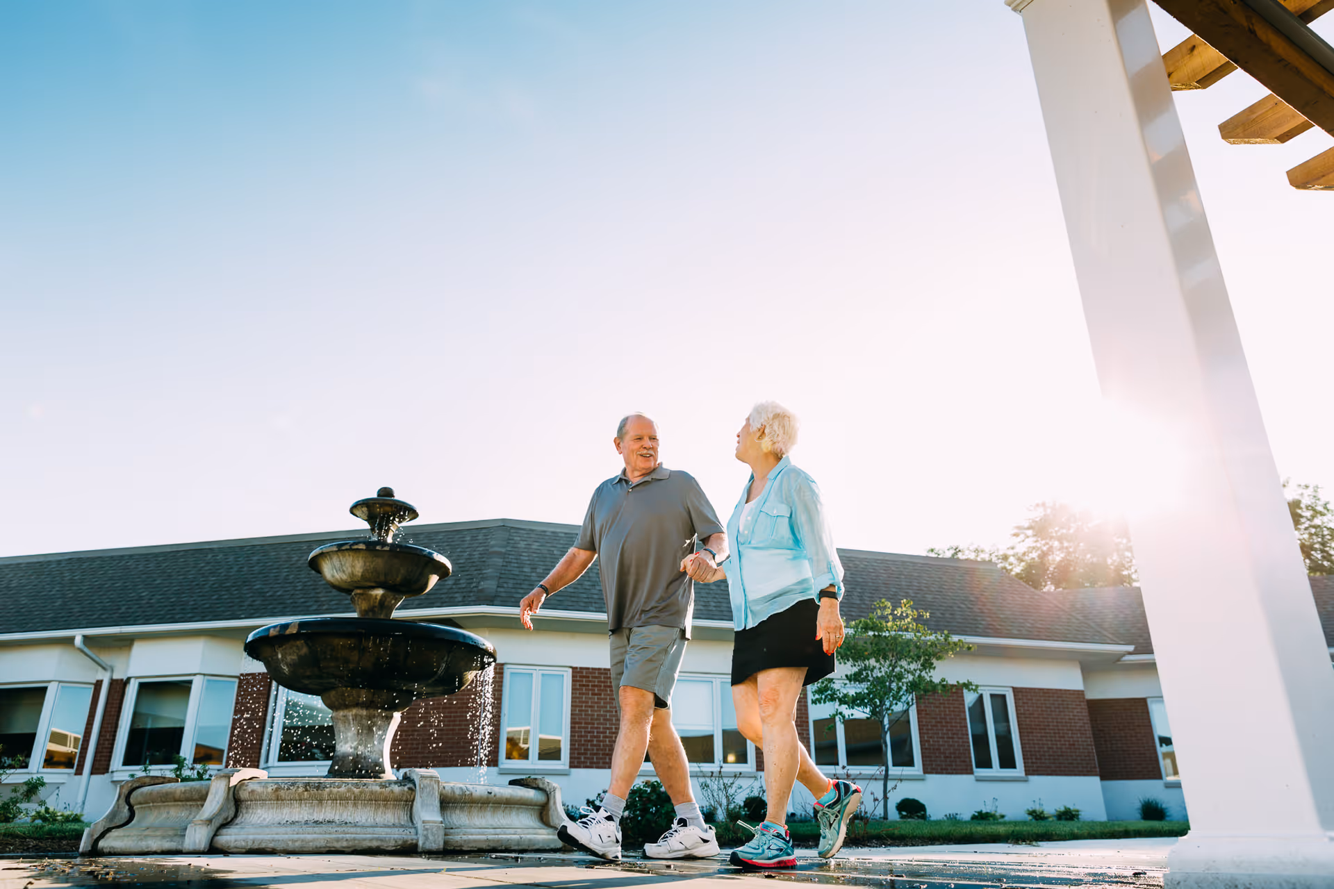 Two seniors walking hand-in-hand past a fountain outside a single-story building at sunset.
