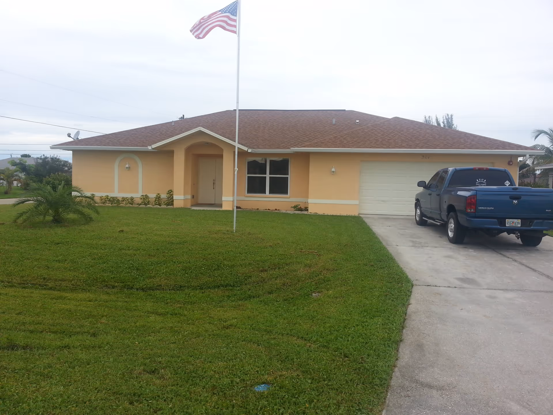 Single-story peach-colored house with a brown roof, a front lawn with a small palm tree, an American flag on a flagpole, and a blue pickup truck parked in the driveway.