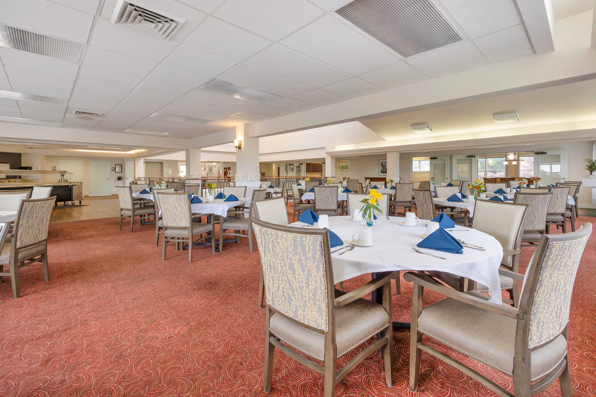 A spacious dining room with multiple round tables covered with white tablecloths, each set with blue folded napkins, white cups, and silverware. The room has patterned carpet flooring, cushioned chairs, and soft lighting from ceiling fixtures. Large windows and mirrors are visible in the background.