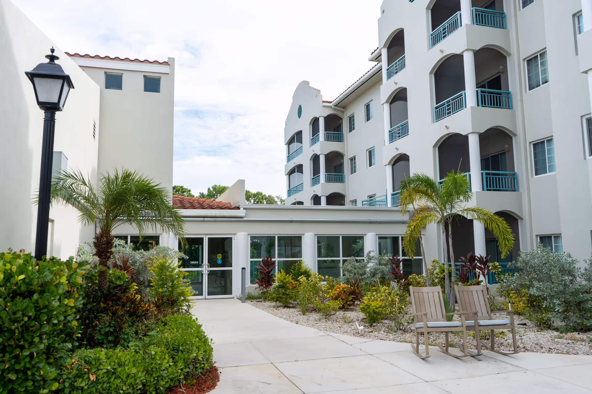 Exterior view of Heron Club at Prestancia showing a multi-story building with balconies, a pathway leading to glass double doors, landscaped greenery including palm trees and bushes, and two wooden rocking chairs on the right side of the path.