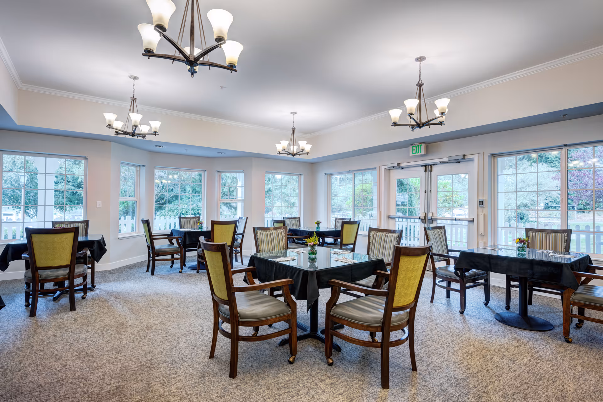 A bright dining room with multiple tables covered in black tablecloths, each surrounded by wooden chairs with cushioned seats and backs. Large windows line the walls, allowing natural light to fill the room and offering views of greenery outside. Elegant chandeliers hang from the ceiling, and small flower arrangements are placed on each table.