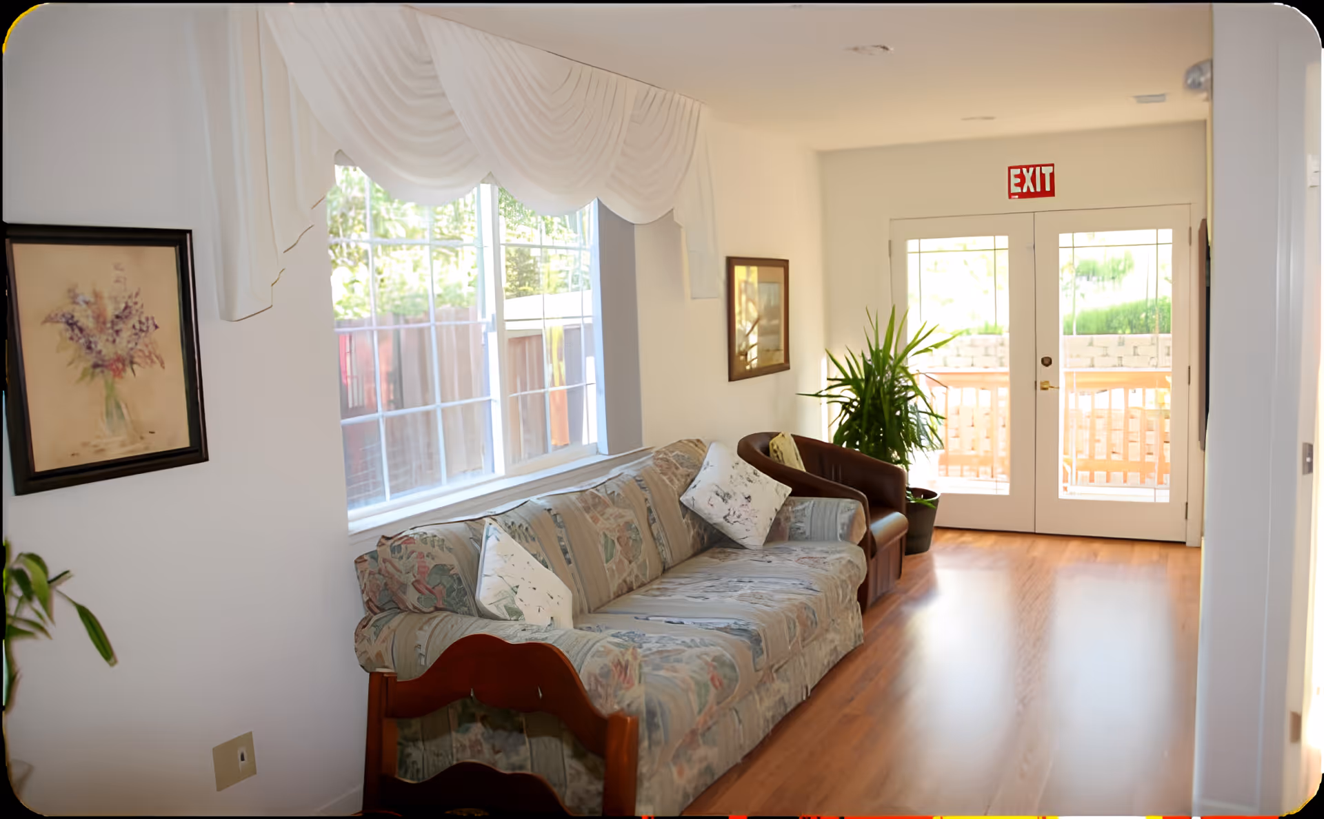 A bright and cozy living room area with a floral patterned sofa and a brown armchair. The room has wooden flooring, a large window with white drapes, a framed floral painting on the wall, a potted plant near glass double doors with an exit sign above, and a wooden chair partially visible in the foreground.