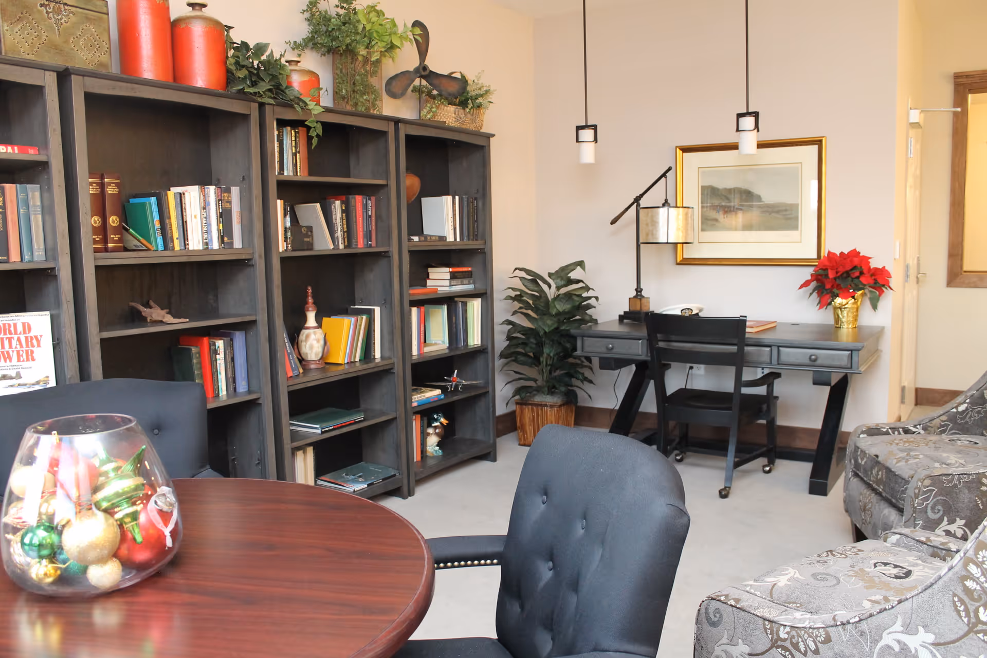 A cozy interior room featuring a round wooden table with a glass bowl filled with colorful ornaments, two upholstered chairs, a large dark wood bookshelf filled with books and decorative items, a black desk with a chair, a potted plant, a framed landscape painting on the wall, and a patterned armchair.