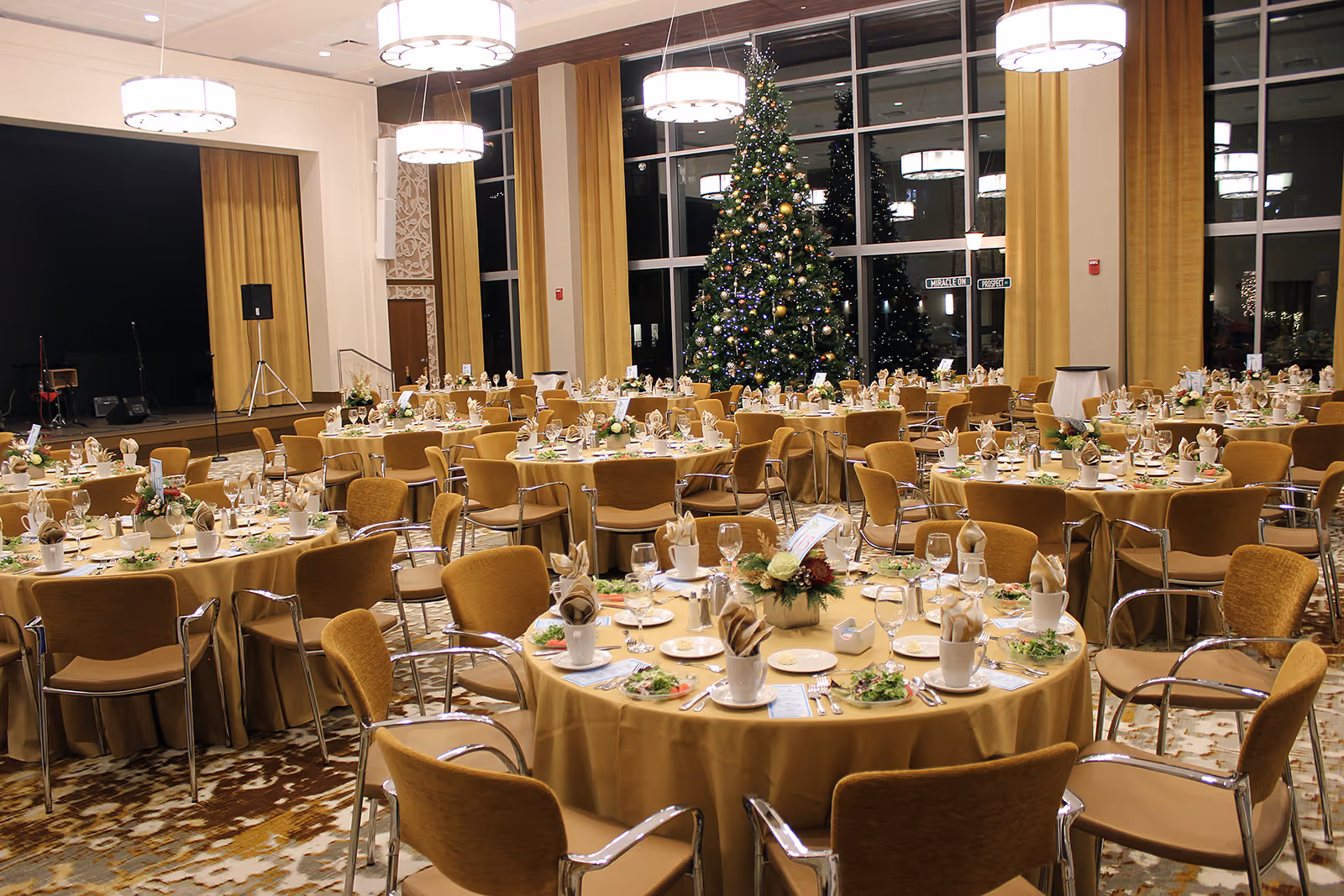 A large dining room decorated for a formal event with round tables covered in beige tablecloths, set with plates, glasses, napkins, and floral centerpieces. A decorated Christmas tree stands near large windows with gold curtains, and a stage with speakers is visible in the background.