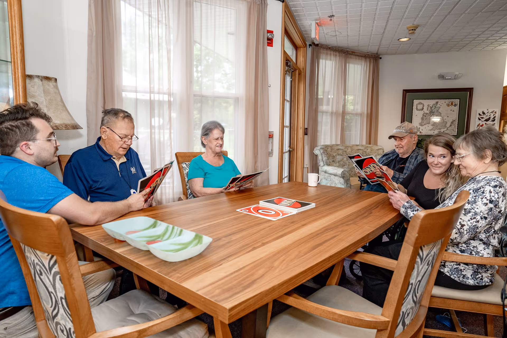 A group of six people, including elderly individuals and a younger person, sitting around a wooden table in a well-lit room with large windows and sheer curtains. They are holding UNO cards and appear to be engaged in a game. The room has comfortable chairs, a lamp, and framed artwork on the walls.