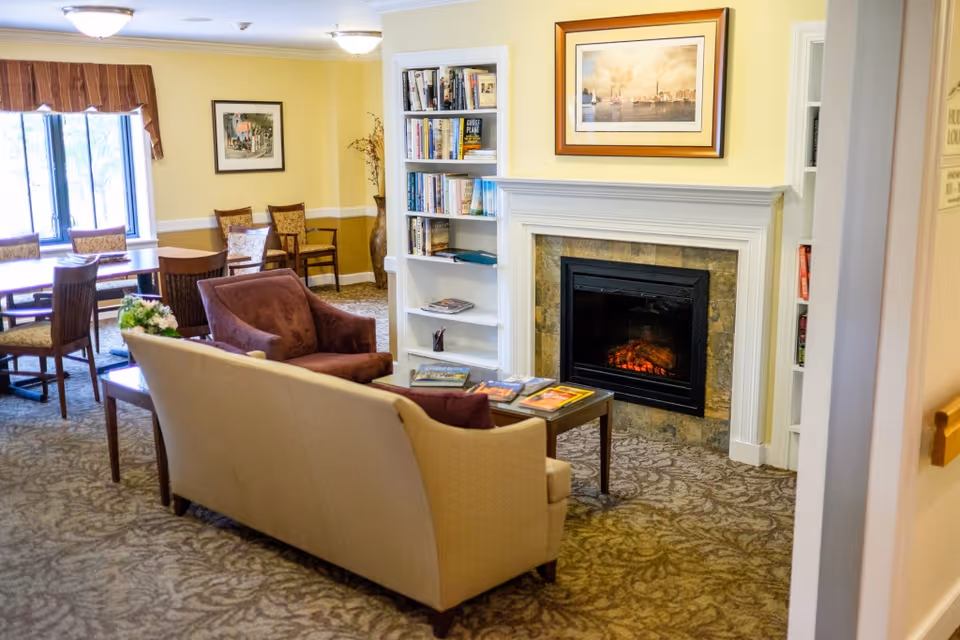 Cozy communal living room with sofas and armchairs facing a fireplace, a bookshelf, and a dining table by the window.