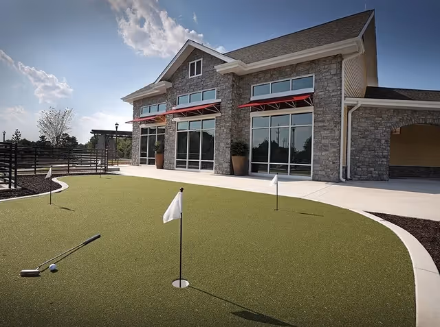 Outdoor putting green with three small flags and a golf club and ball on the green, in front of a stone building with large windows and red awnings under a partly cloudy sky.