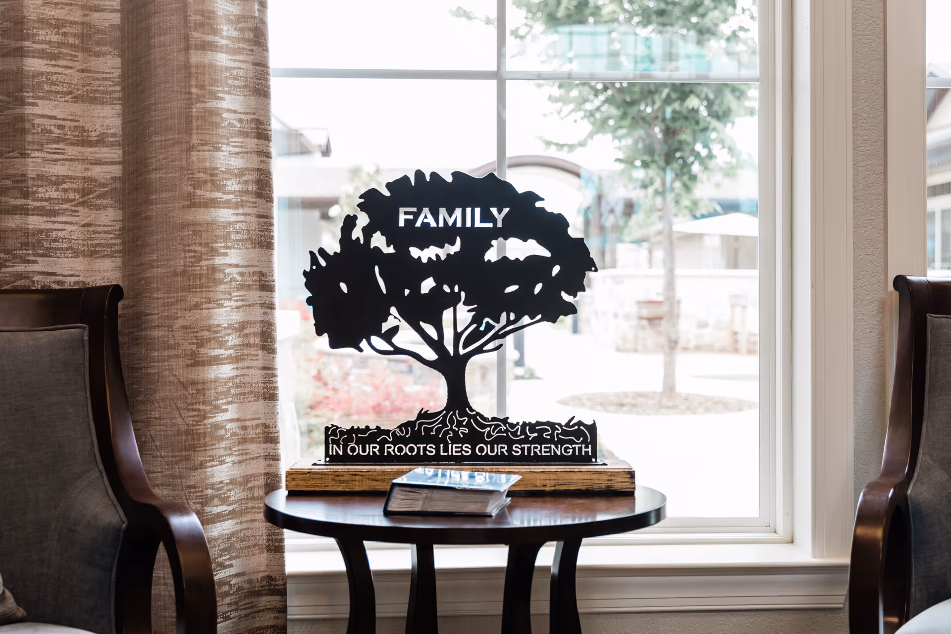 A decorative metal tree sculpture with the word 'FAMILY' at the top and the phrase 'IN OUR ROOTS LIES OUR STRENGTH' at the base, placed on a round wooden table in front of a large window. Two upholstered chairs flank the table, and beige curtains are partially visible on the left side.