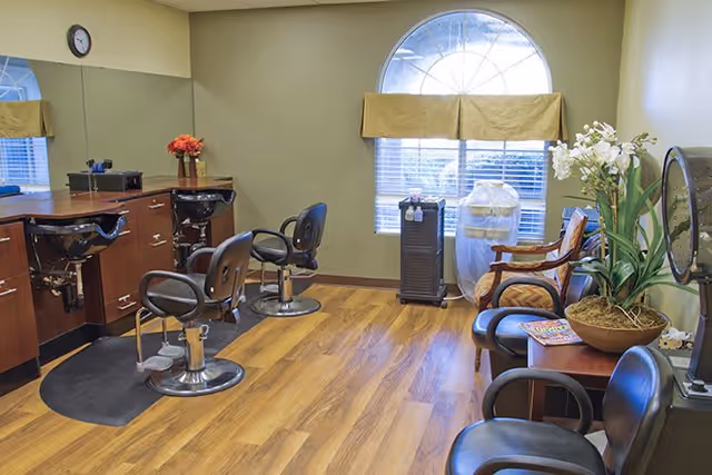 Interior of a salon area with two black salon chairs in front of a wooden counter with sinks, a large mirror on the wall, a clock, and a window with a half-circle top letting in natural light. There are additional chairs and a small table with magazines and a potted plant in the waiting area.