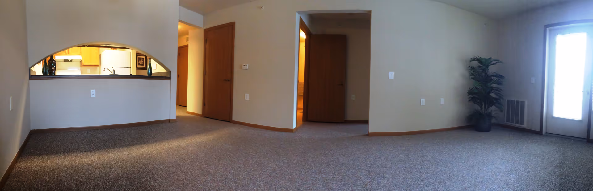 Empty living room with beige walls and carpeted floor, a large window or door letting in natural light on the right, a potted plant in the corner, and an open archway showing a kitchen area with wooden cabinets and white appliances. There are two wooden doors visible in the middle of the image.