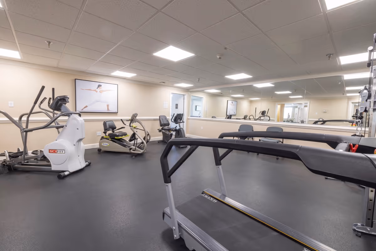 A fitness room in Heatherwood Retirement Community featuring exercise equipment including a treadmill, elliptical machine, and stationary bikes. The room has a large mirror on one wall, bright overhead lighting, and a framed picture of a person doing a yoga pose on the wall.