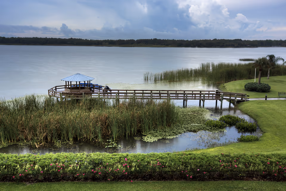 A wooden pier with a small covered gazebo extends over a lake surrounded by reeds and manicured lawns.