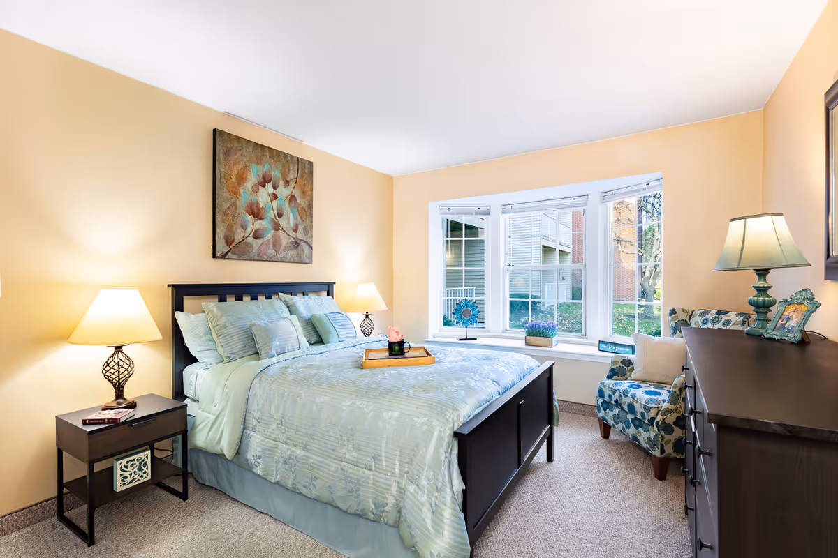 A bright and cozy bedroom with a large bed dressed in light blue bedding and multiple pillows. Two bedside tables each hold a lamp, and a decorative painting hangs above the bed. A bay window with white blinds lets in natural light, and a patterned armchair with a pillow sits next to a dark wooden dresser with a lamp and framed photo on top.
