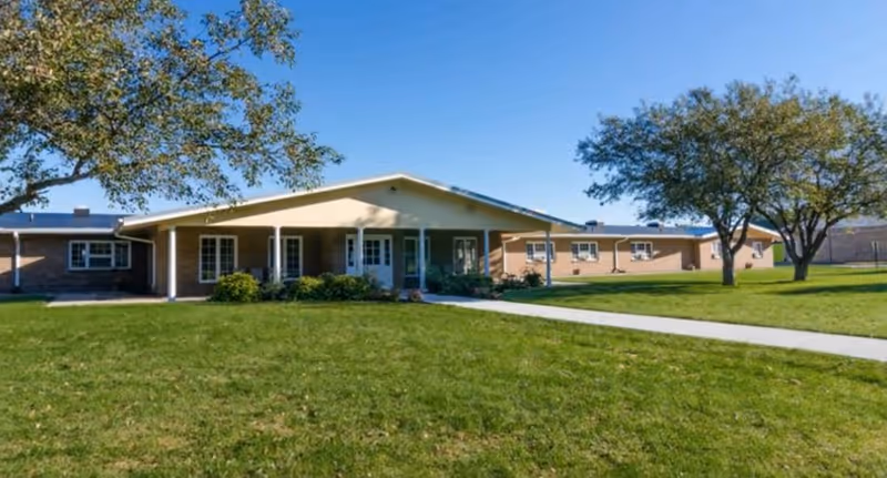 Single-story healthcare building with a covered entrance, a wide front lawn, sidewalk, and trees under a clear blue sky.