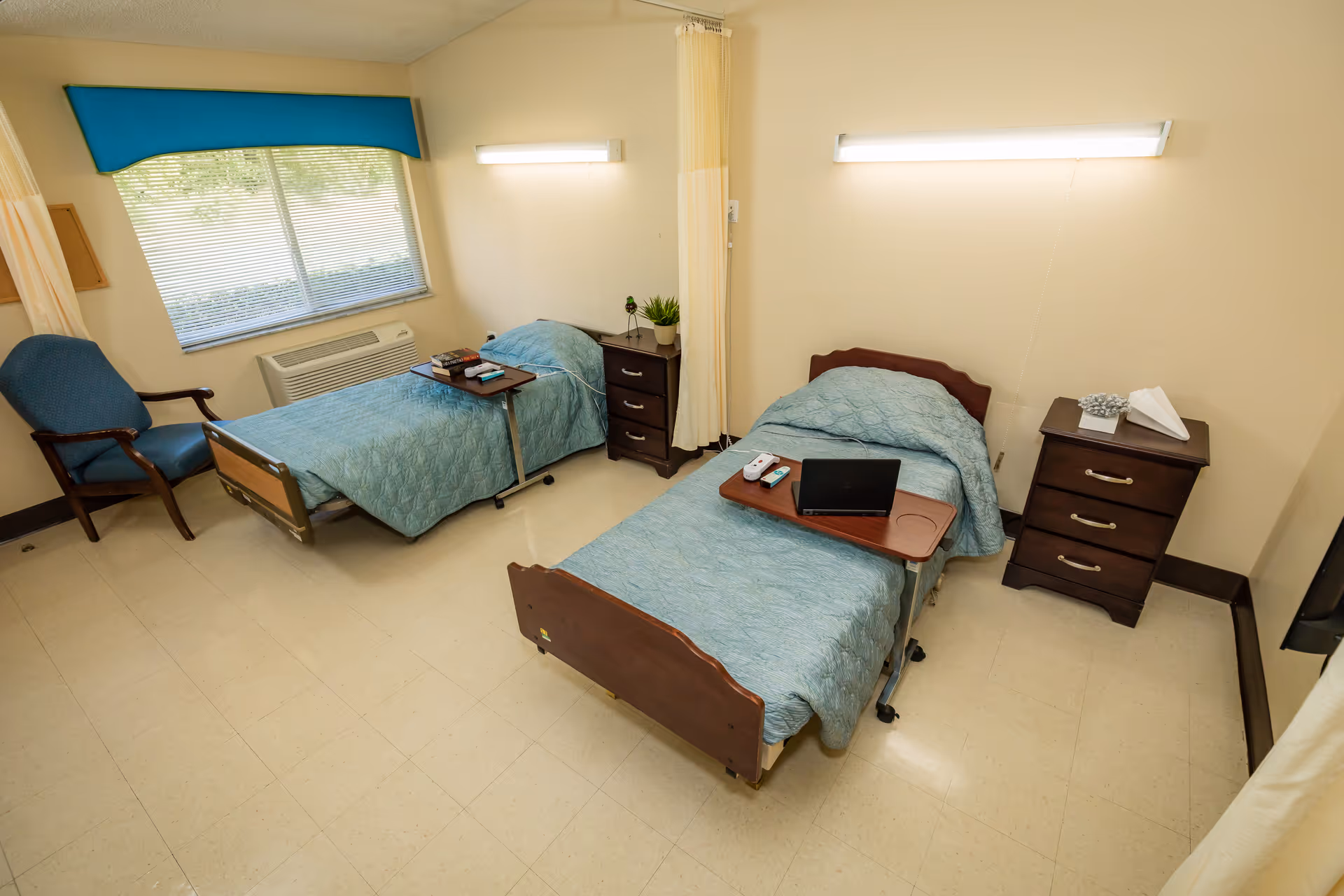 A room with two single hospital-style beds each covered with a blue quilt. Each bed has a small overbed table with items on it, including a laptop, remote controls, and books. There are two dark wooden nightstands, one between the beds and one beside the right bed, with a plant and tissues on top. A blue armchair is positioned near a window with blinds and a blue valance. The room has beige walls and a tiled floor, with a curtain divider between the beds.