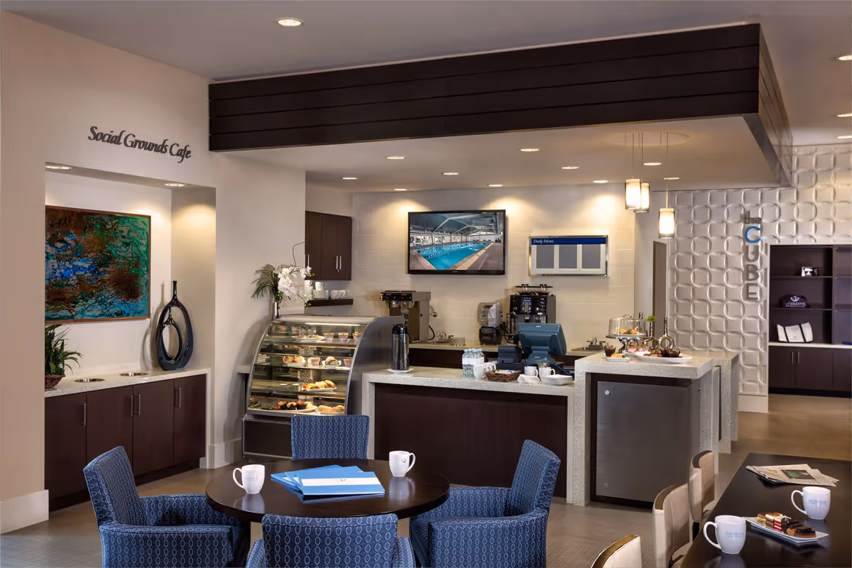 Interior view of a modern cafe area named Social Grounds Cafe with a round table surrounded by blue patterned chairs, a counter displaying pastries, coffee machines, and a TV screen showing a swimming pool. The space has warm lighting, decorative wall art, and a sign reading 'the CUBE' on a textured white wall.