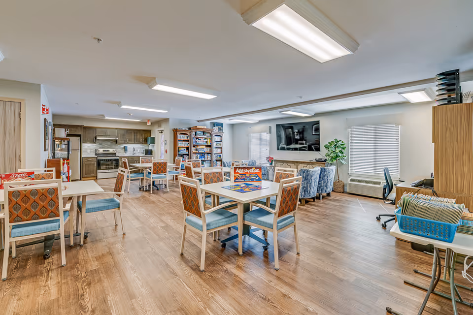 A spacious common area in a senior living facility featuring several tables and chairs arranged for group activities or dining. In the background, there is a kitchen area with stainless steel appliances, including a refrigerator and stove. A large flat-screen TV is mounted on the wall above a fireplace, with comfortable armchairs arranged nearby. Shelves filled with board games are visible, and a desk with office supplies is positioned near a window with blinds. The room has wood flooring and bright overhead lighting.