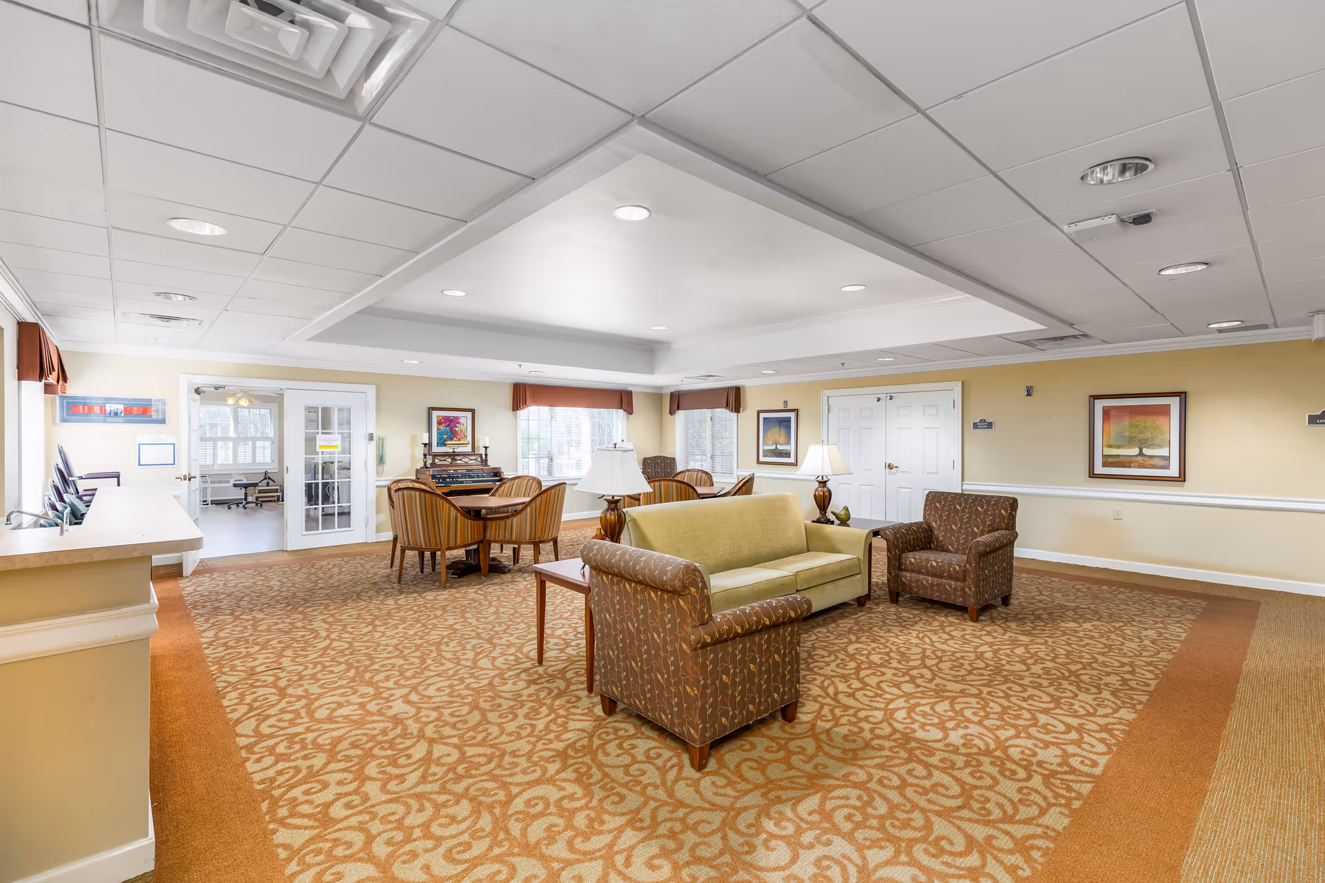 A spacious senior living facility common area with patterned carpet, a green sofa, two patterned armchairs, wooden side tables with lamps, and a dining table with four striped chairs. The room has beige walls adorned with framed artwork, large windows with valances, and a white ceiling with recessed lighting. A double door and a glass door leading to another room are visible.