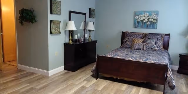 A bedroom with a wooden bed frame and a bed made with patterned bedding and multiple pillows. There is a dark wooden dresser with two lamps and a mirror above it. The walls are light blue with decorative wall art, and the floor is light wood. A doorway leads to another room, and there is a small plant hanging on the wall near the doorway.