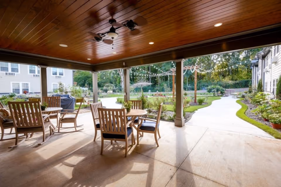 Covered outdoor patio area with wooden ceiling and ceiling fans, furnished with round wooden tables and chairs. Beyond the patio is a paved walkway surrounded by landscaped gardens and greenery, with string lights hanging overhead.