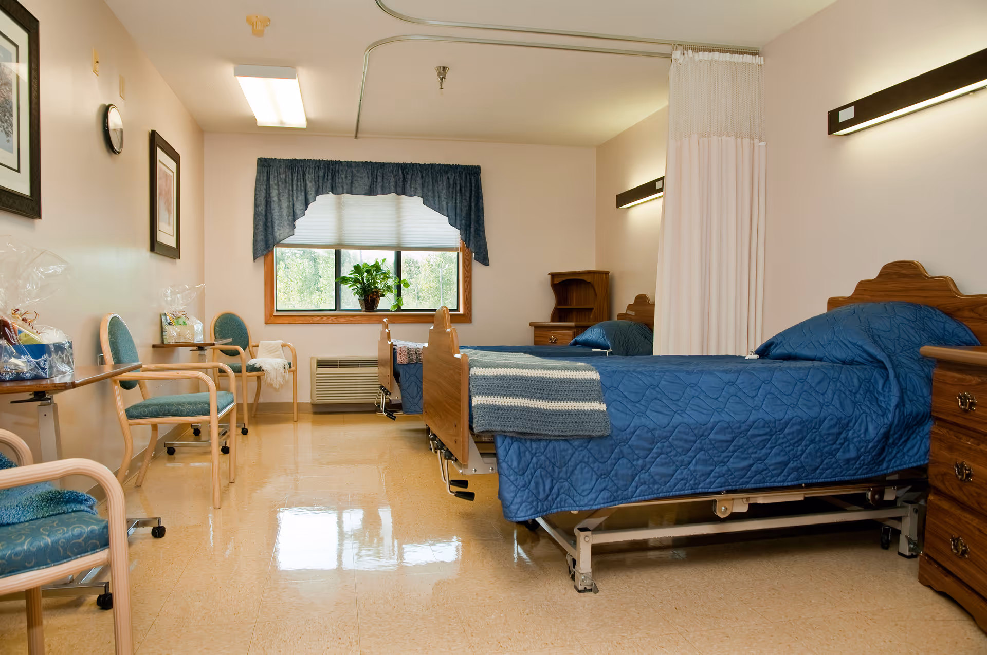 A clean and well-lit senior care facility bedroom with two hospital-style beds covered in blue quilts. The room has a window with blue curtains and a potted plant on the windowsill. There are several chairs along the left wall, a wooden nightstand with drawers, and a privacy curtain partially drawn between the beds. The floor is shiny and reflective, and the walls are decorated with framed artwork.