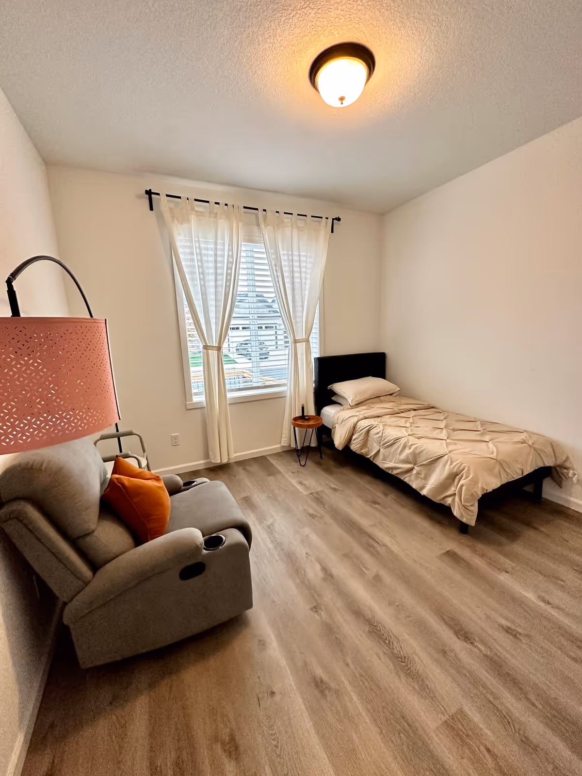 A simple bedroom with a single bed covered with a beige comforter and a pillow, a small round wooden side table next to the bed, a gray recliner chair with an orange pillow, a pink floor lamp, and a window with white curtains letting in natural light.