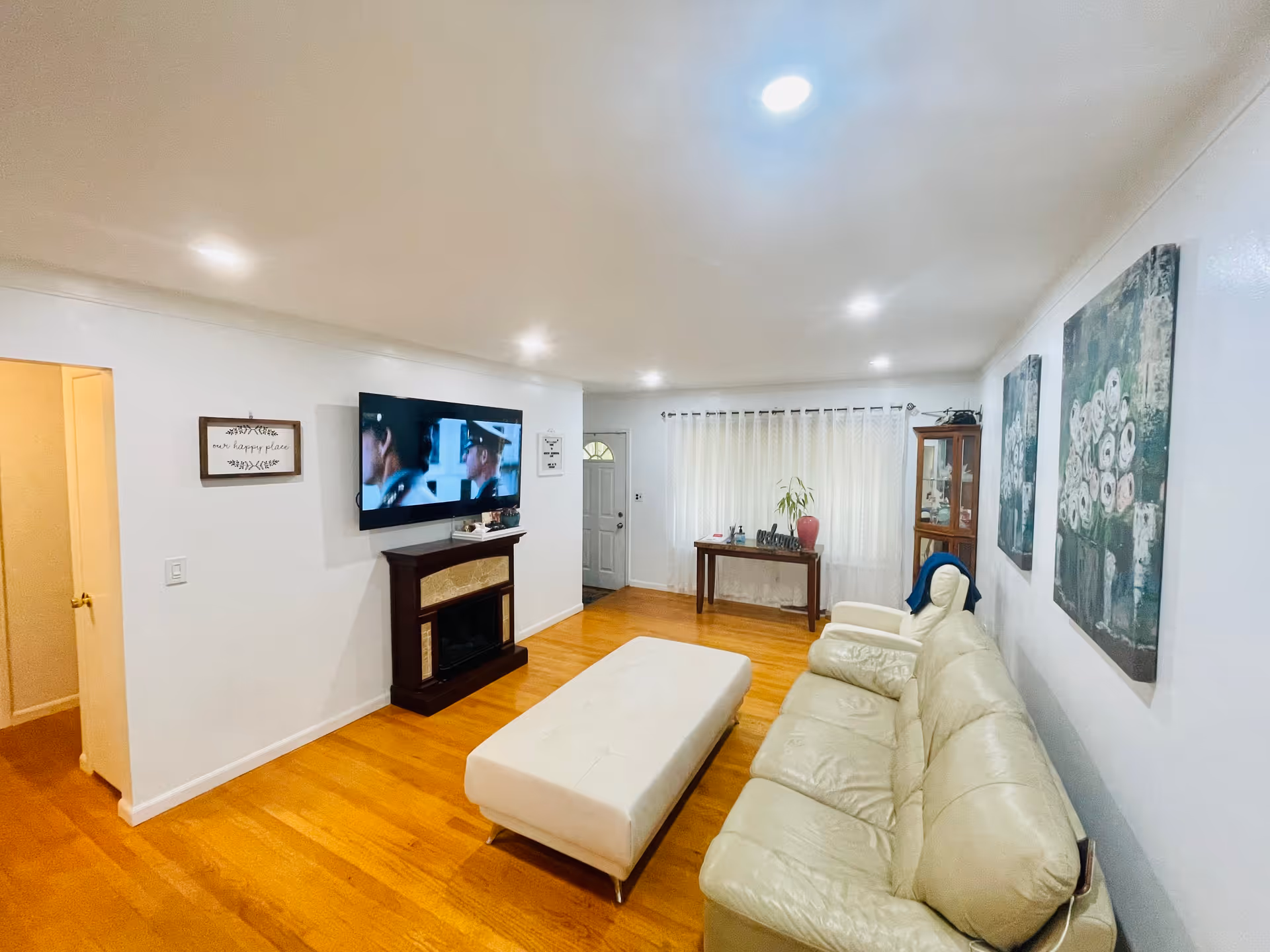 Bright living room with a cream sofa and ottoman, wall-mounted TV above a fireplace, hardwood floors and a console table by a curtained window.
