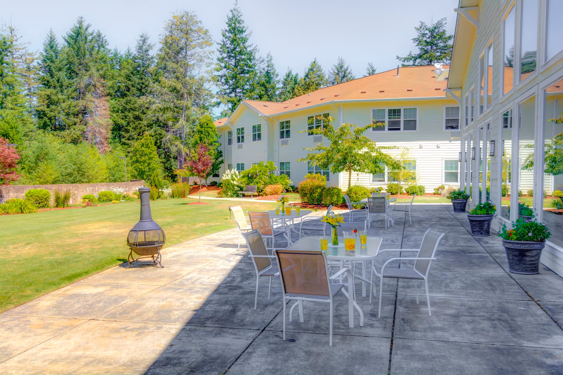 Outdoor patio area at Ocean Ridge by Cogir with several white tables and chairs arranged on a concrete surface. The tables have drinks and a vase with yellow flowers. There are large windows on the right side of the building, potted plants along the patio, and a grassy lawn with trees and shrubs in the background under a clear sky.
