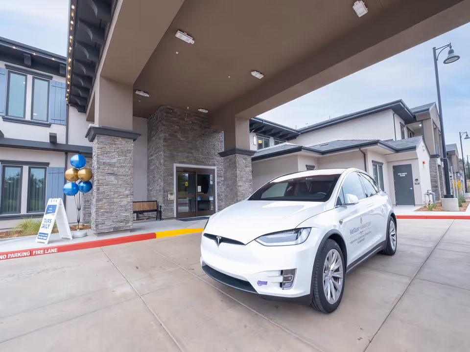 Front entrance of a senior living facility with a covered drop-off, a white Tesla parked in front, and balloons by the doorway.