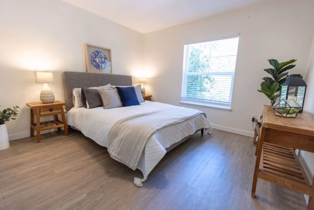 A bright bedroom with a large bed featuring a gray upholstered headboard and white bedding with multiple pillows in shades of gray and blue. There are two wooden nightstands on either side of the bed, each with a lamp. A framed artwork hangs above the bed. A window with white blinds lets in natural light. On the right side of the room, there is a wooden console table with decorative items and a green potted plant. The floor is light wood, and the walls are painted white.