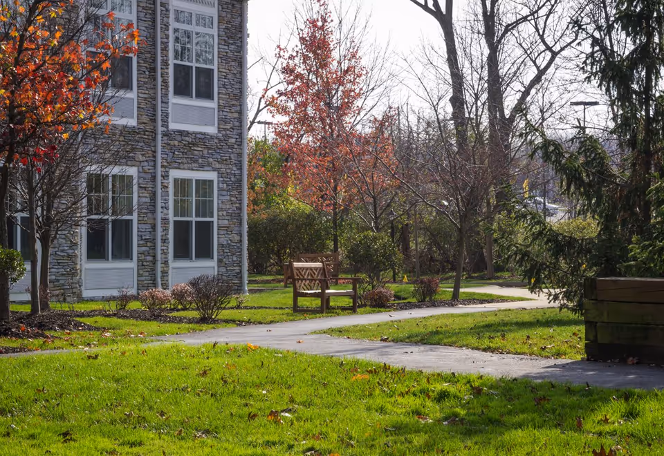 Outdoor garden area at Sunrise at Parma featuring a stone building facade with multiple windows, a paved walking path, wooden benches, green grass, and trees with autumn foliage.