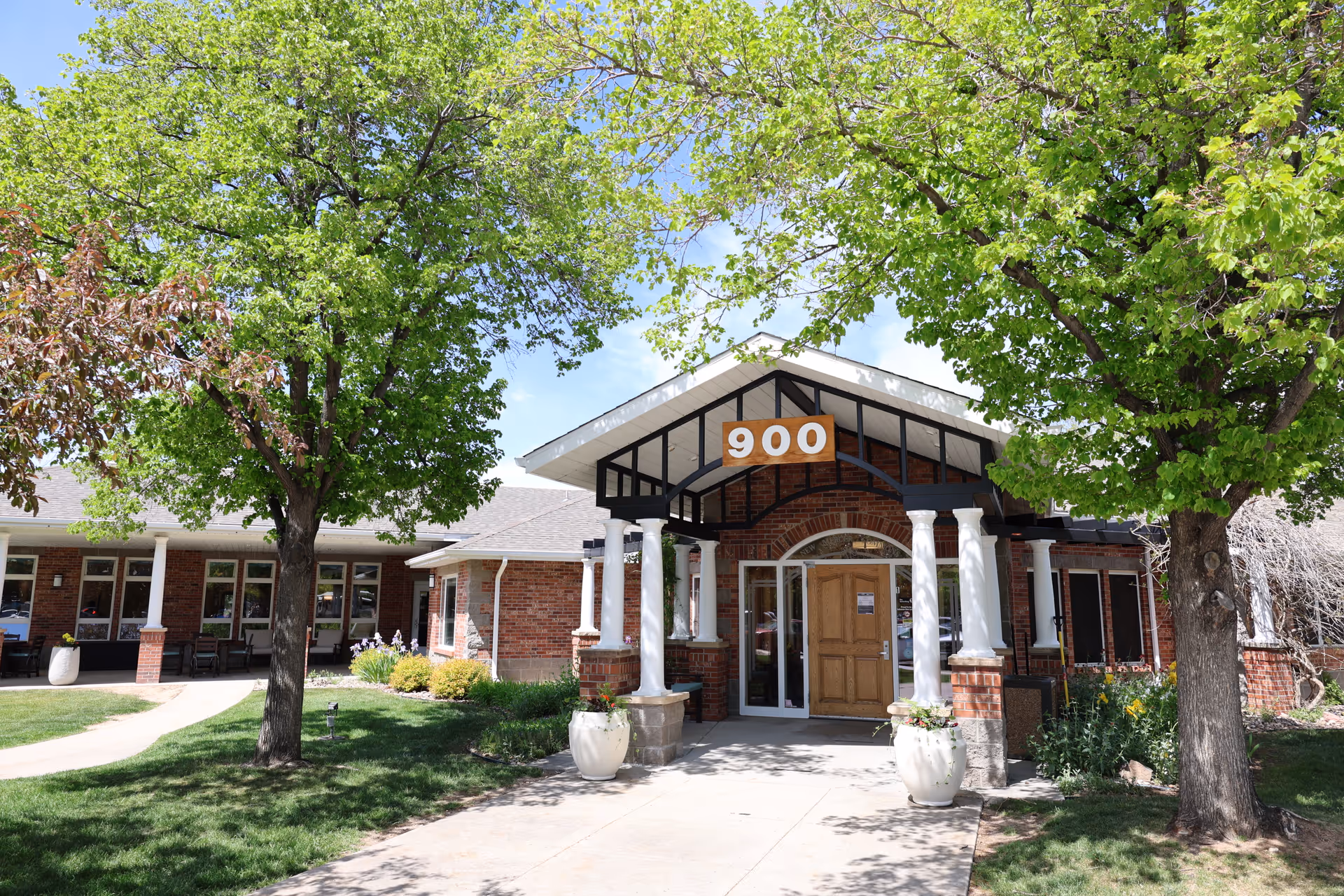 Front entrance of a brick assisted living building with white columns, a wooden door under a '900' sign, walkway and green trees.