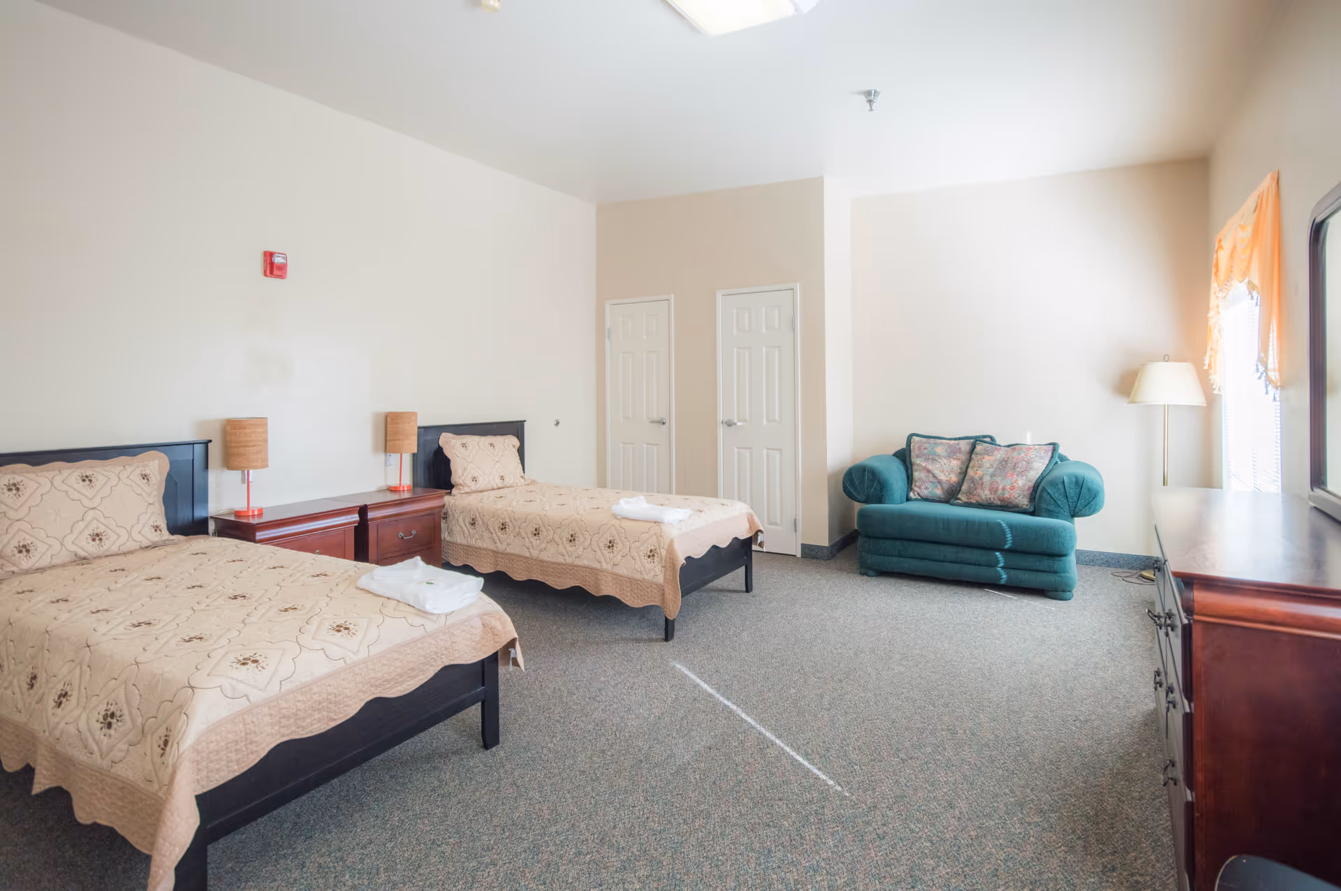 A bright and spacious bedroom in an assisted living community featuring two twin beds with beige patterned bedspreads, a wooden nightstand with two lamps between the beds, a green upholstered loveseat with floral cushions, a floor lamp, and a wooden dresser with a mirror. The room has light-colored walls, carpeted flooring, and two closed white doors.