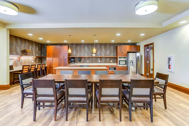 A spacious, modern kitchen and dining area with a long wooden dining table surrounded by ten chairs. The kitchen features wooden cabinets, a stainless steel refrigerator, built-in oven, microwave, and a kitchen island with three pendant lights hanging above. There is also a counter with four high chairs along the left wall. The room has light-colored walls, wood flooring, and recessed ceiling lights.