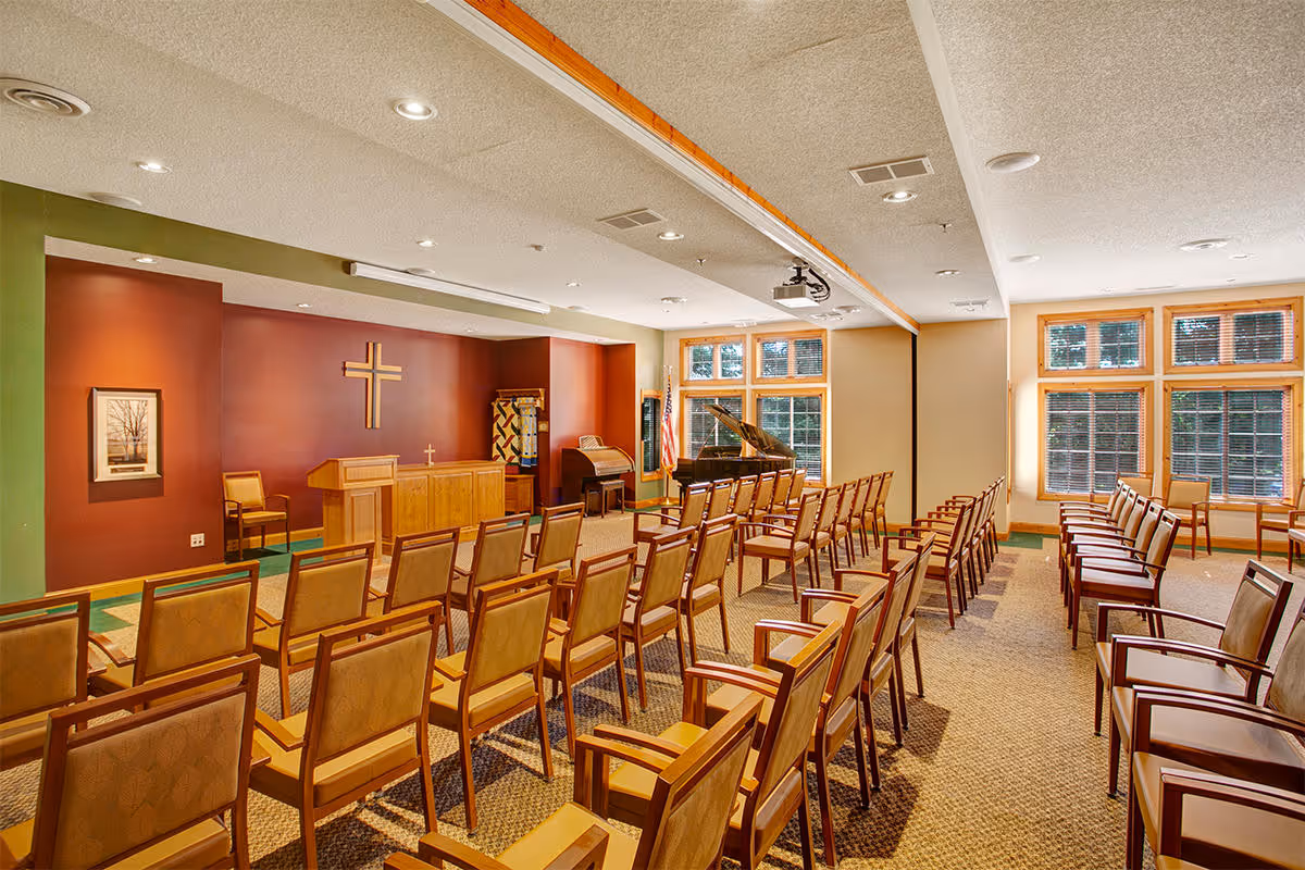 A chapel or meeting room with rows of wooden chairs facing a wooden podium and a wall-mounted cross. The room has large windows with wooden frames letting in natural light, a piano in the corner, and a carpeted floor.