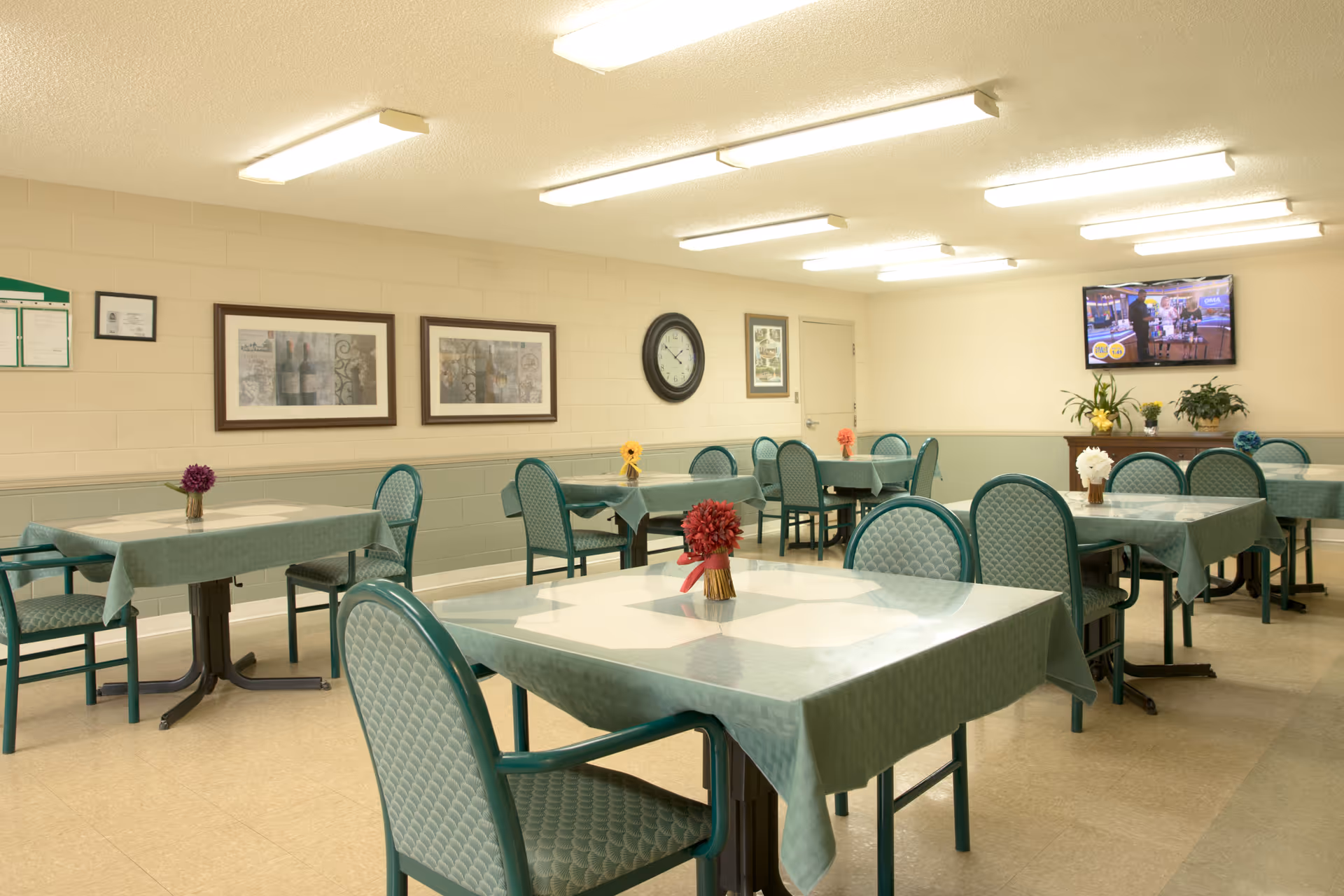 A dining room with several tables covered in green tablecloths, each table decorated with a small flower arrangement. Green cushioned chairs surround the tables. The walls are light-colored with framed pictures and a clock. A television is mounted on the far wall above a cabinet with plants.