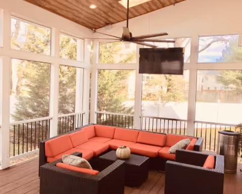 Screened outdoor seating area with red-cushioned wicker sectional, ceiling fan, wall-mounted TV, and a small table with a decorative pumpkin.