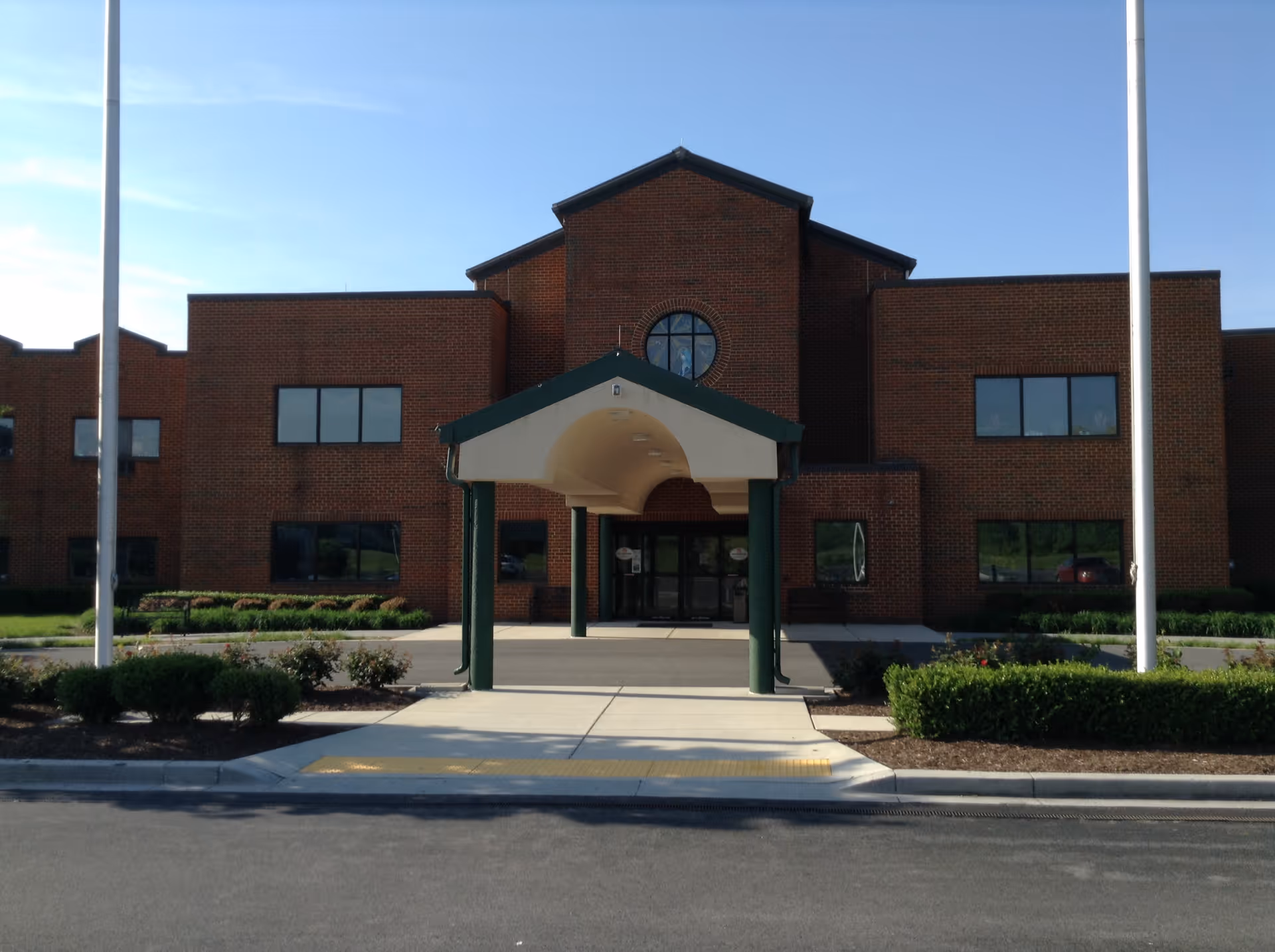 Front exterior view of a brick building with a covered entrance supported by green pillars, two flagpoles on either side, and landscaped bushes in front under a clear blue sky.